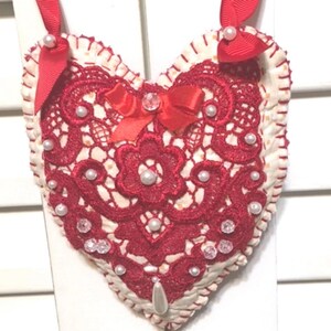 May include: Red heart-shaped ornament with white lace and pearl embellishments. A red satin bow with a rhinestone center is attached to the center of the heart.