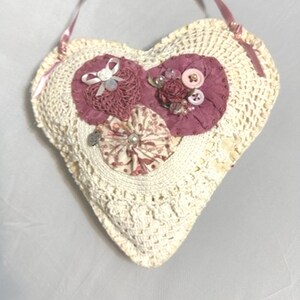 May include: A white crocheted heart-shaped pillow with pink fabric flowers and buttons.
