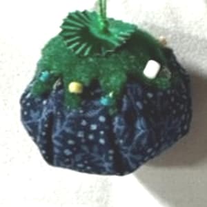 May include: A blue and green pincushion with a floral pattern. The pincushion is round and has a green felt top with a decorative pinwheel.