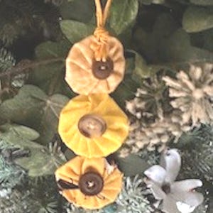 May include: Three handmade fabric ornaments in shades of yellow and tan, each with a dark brown button center. The ornaments are suspended by a yellow string, and are displayed against a backdrop of greenery and white flowers. These are decorative holiday ornaments.