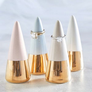 May include: Four ceramic ring holders in the shape of cones. The cones are two-toned, with a light blue, white, or pink top and a gold bottom. Each cone has a ring on top.