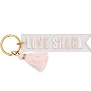 Puede incluir: Un llavero de acrílico transparente con un borla rosa. El llavero tiene las palabras "LOVE SHACK" escritas en letras blancas con un contorno rosa.