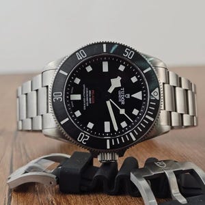 May include: A Tudor Pelagos wristwatch with a black dial, white luminescent hour markers, and a rotating bezel. The watch features a brushed titanium link bracelet and is accompanied by a separate black rubber strap with a metal buckle.