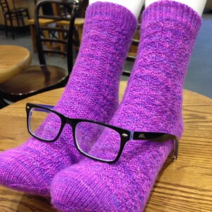 May include: A pair of purple and pink knitted socks with a pair of black framed glasses resting on top.