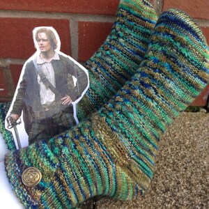 Op de afbeelding: Handgebreide sokken in groen, blauw en bruin met een textuurpatroon en een decoratieve knoop. Een uitgesneden afbeelding van een man in een kilt staat op de achtergrond.