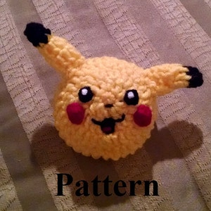 Könnte beinhalten: Eine gehäkelte gelbe Pikachu-Figur mit schwarzen Ohren und Augen, roten Wangen und einem schwarzen Lächeln. Die Figur sitzt auf einem gestreiften Stoffhintergrund.