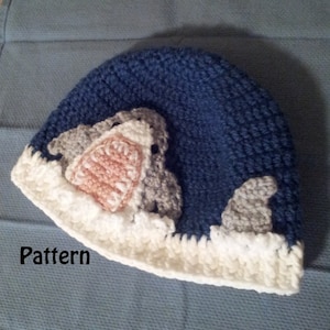 May include: A handmade blue crochet hat with a shark design. The hat features a white brim and a shark with gray, pink, and white details. The word "Pattern" is printed on the lower left side of the image.