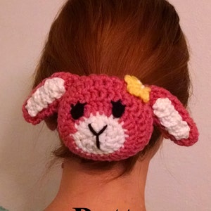 May include: A pink and white crocheted bunny hair scrunchie with black eyes and a yellow flower. The scrunchie is worn in a woman's hair.