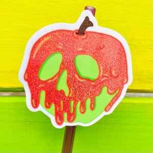 May include: A glittery red apple-shaped decoration with a green skull design. The apple has a brown stem and a white border. The decoration is on a brown stick, set against a yellow and green background.