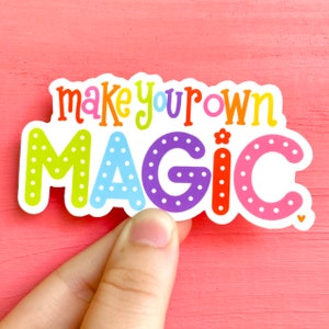 May include: A colorful sticker with the words "make your own MAGIC" in various bright colors. The sticker has a white border and is held by a hand against a pink background.