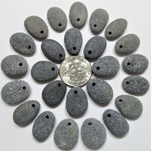 May include: A collection of 20 smooth, gray river rocks with holes drilled through them. The rocks are arranged in a circle around a US dime.