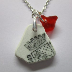 Könnte beinhalten: Eine Halskette mit einem weißen Keramik-Anhänger, der ein schwarz-weißes Bild eines Wappens mit einer Krone und einem Schild zeigt. Ein roter Glasanhänger hängt an dem Anhänger.