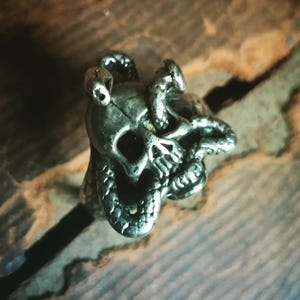 May include: A silver-toned ring featuring a skull design with snakes coiled around it. The skull has detailed features, and the snakes add a textured element. The ring is likely made of metal and has a gothic or biker aesthetic.