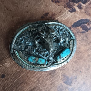 Peut inclure: Boucle de ceinture ovale argentée avec une tête de bélier au centre, flanquée de pierres turquoise. La boucle présente des volutes et des motifs de feuilles complexes, créant une esthétique rustique.