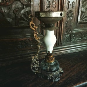 Puede incluir: Una lámpara de aceite antigua con una chimenea de vidrio blanco y una base de metal decorativa. La lámpara tiene una parte superior de metal dorado con la inscripción "Patentado 1905, U.S.A."