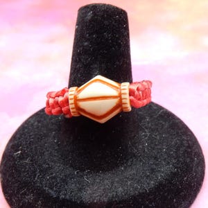 以下が含まれることがあります： 赤色の紐のブレスレットに、白とオレンジの幾何学模様のビーズが付いている。ビーズは丸みを帯びた角を持つダイヤモンドの形をしている。