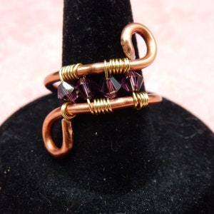 May include: A copper wire ring with a gold wire wrap and purple crystals. The ring is on a black display stand.