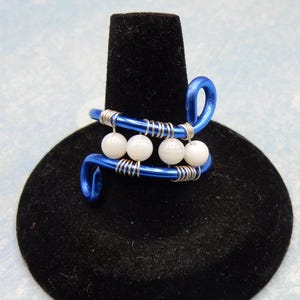 May include: A blue wire ring with four white pearl beads. The ring is on a black display stand.