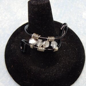 Puede incluir: Un anillo de alambre negro con cuentas de cristal transparente. El anillo se muestra en un soporte de anillo negro.