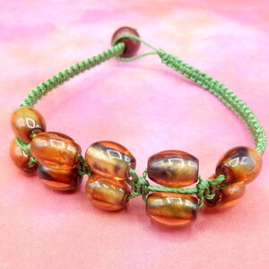 May include: A handmade bracelet featuring amber-colored, oval-shaped beads with dark accents. The beads are strung on a green, braided cord. A single, round, dark brown bead is used as a clasp. The bracelet is displayed on a pink background.