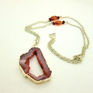 May include: A necklace featuring a large, irregular, reddish-brown geode slice pendant with a gold-toned metallic edge. The pendant hangs from a thin silver-toned metal chain that includes two small, smooth, orange-brown oval beads.