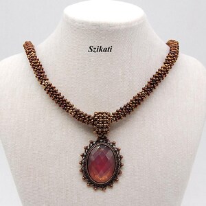 May include: A beaded necklace with a large, oval pendant. The pendant features a faceted, reddish-orange stone set in a decorative, bronze-coloured frame. The necklace is displayed on a white mannequin and has the text "Szikati".
