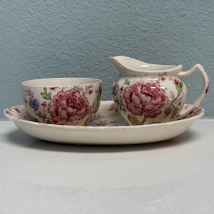May include: A cream-colored ceramic creamer and a matching bowl resting on an oval tray. Each piece features a repeating pattern of large pink flowers with green leaves and small blue accents. The set has a smooth, glossy finish and scalloped edges.