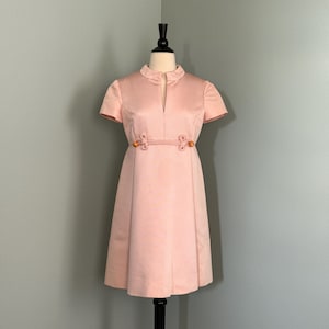 Puede incluir: Un vestido vintage rosa con mangas cortas y cuello mandarín. El vestido tiene un cinturón anudado con botones de madera.