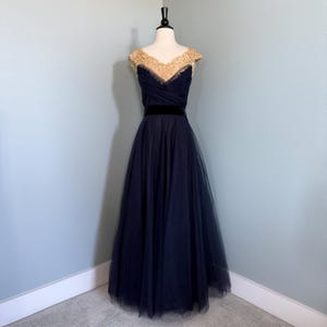 Może przedstawiać: Granatowa suknia wieczorowa ze złotym, koralikowym stanikiem i czarnym aksamitnym paskiem. Długa spódnica wykonana jest z lejącej tkaniny tiulowej. Suknia jest prezentowana na manekinie na tle jasnoniebieskiej ściany.