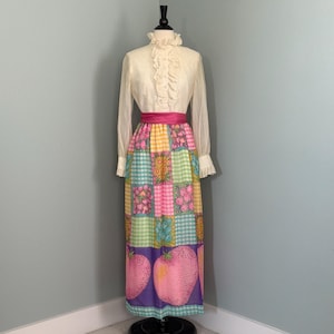 Puede incluir: Un vestido vintage color crema con cuello alto con volantes y mangas largas. El vestido presenta una falda patchwork con estampados florales y frutales en rosa, amarillo, verde y azul, y una faja rosa brillante. El vestido se exhibe en un maniquí.