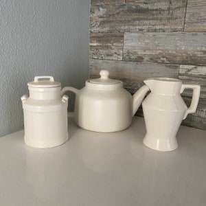 Peut inclure: Trois articles de cuisine en céramique blanche : une théière, un pichet et un bidon à lait. La théière a un corps rond et un bec verseur. Le pichet a un corps rond et une anse. Le bidon à lait a un couvercle et une anse.