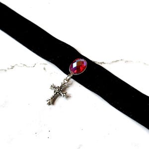 May include: Black velvet choker necklace with a red gemstone pendant and a silver cross charm. The choker is a simple band, and the pendant is an oval-shaped, faceted stone. The cross charm is detailed with floral designs.