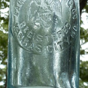 Puede incluir: Una botella de vidrio transparente y antigua con texto en relieve y un emblema circular. El emblema presenta un &aacute;guila con las alas extendidas y las palabras "EAGLE BOTTLING CO. REGISTERED KANSAS CITY MO." La botella tiene un tinte ligeramente azulado.