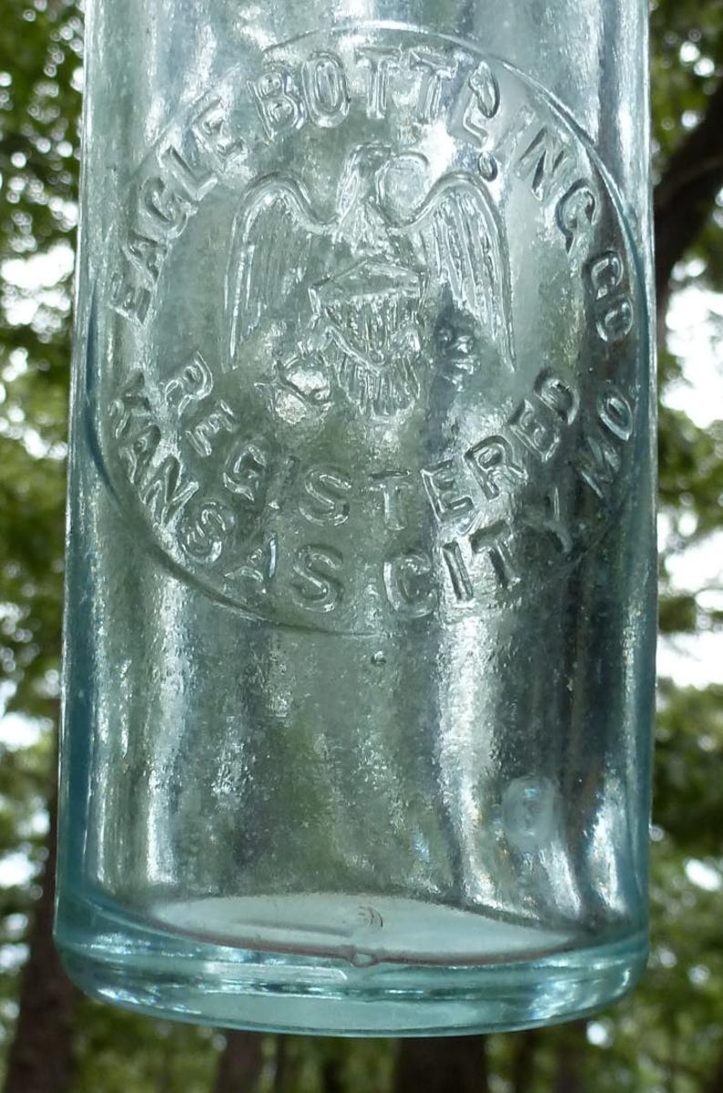 Puede incluir: Una botella de vidrio transparente y antigua con texto en relieve y un emblema circular. El emblema presenta un &aacute;guila con las alas extendidas y las palabras "EAGLE BOTTLING CO. REGISTERED KANSAS CITY MO." La botella tiene un tinte ligeramente azulado.