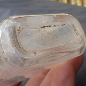 May include: Close-up of the bottom of a clear glass bottle with a slightly frosted finish. The bottle has a rounded rectangular shape.