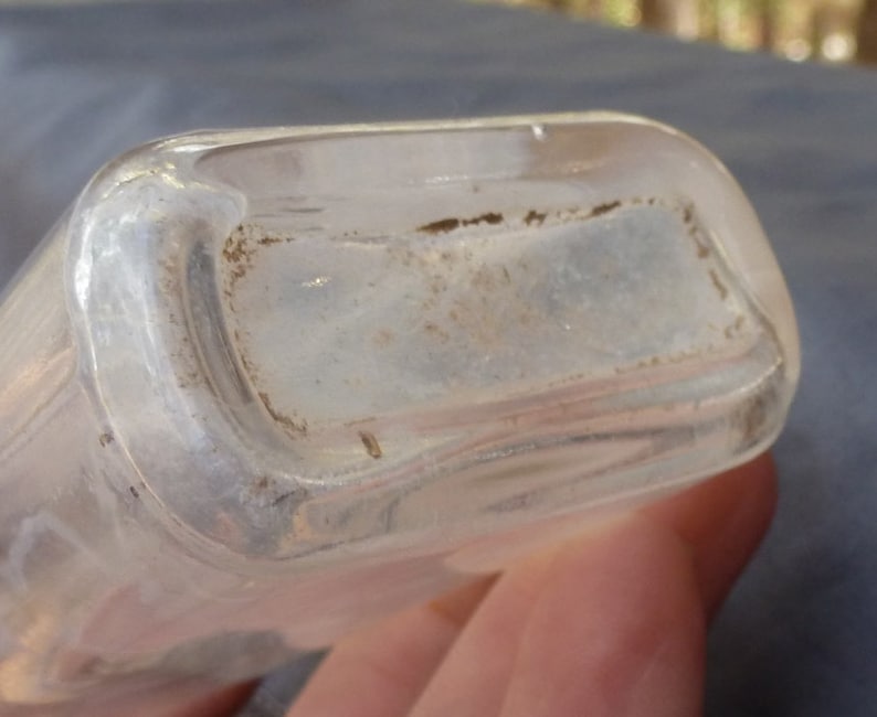 May include: Close-up of the bottom of a clear glass bottle with a slightly frosted finish. The bottle has a rounded rectangular shape.