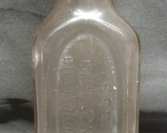 May include: A clear glass bottle with a rounded top and a rectangular body. The bottle has a raised oval on the front with text that is difficult to read.