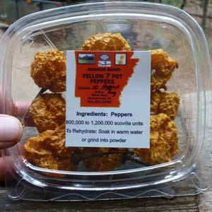 May include: A clear plastic container holds several dried yellow 7 Pot peppers. A label indicates they are Arkansas grown, with a Scoville heat unit range of 800,000 to 1,200,000. The label also includes instructions for rehydration.