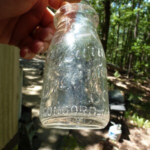 Op de afbeelding: Transparante glazen melkfles met reli&euml;fletters. De fles leest "Cottage Cream Co. Concord, N.H.". De fles wordt omhoog gehouden tegen een achtergrond van bomen en zonlicht.