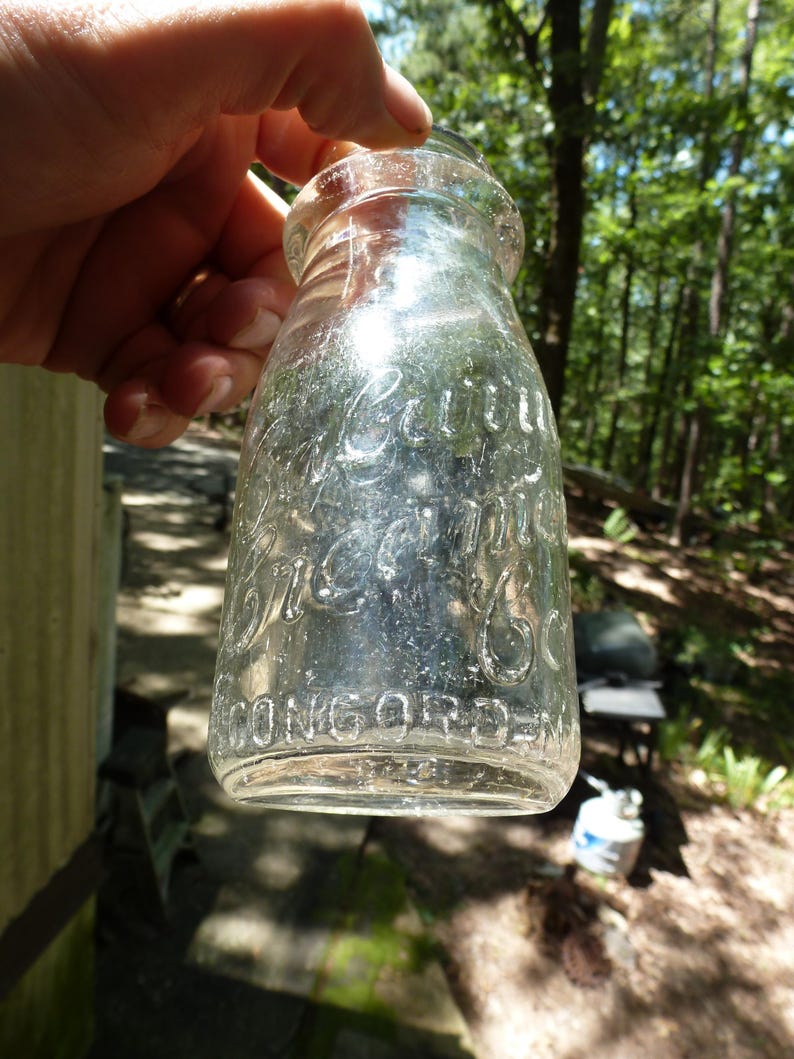 Op de afbeelding: Transparante glazen melkfles met reli&euml;fletters. De fles leest "Cottage Cream Co. Concord, N.H.". De fles wordt omhoog gehouden tegen een achtergrond van bomen en zonlicht.