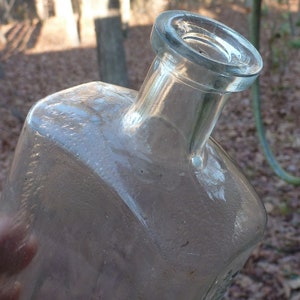May include: A clear glass bottle with a wide mouth and a rounded, slightly flared top. The bottle has a textured surface and appears to be old.