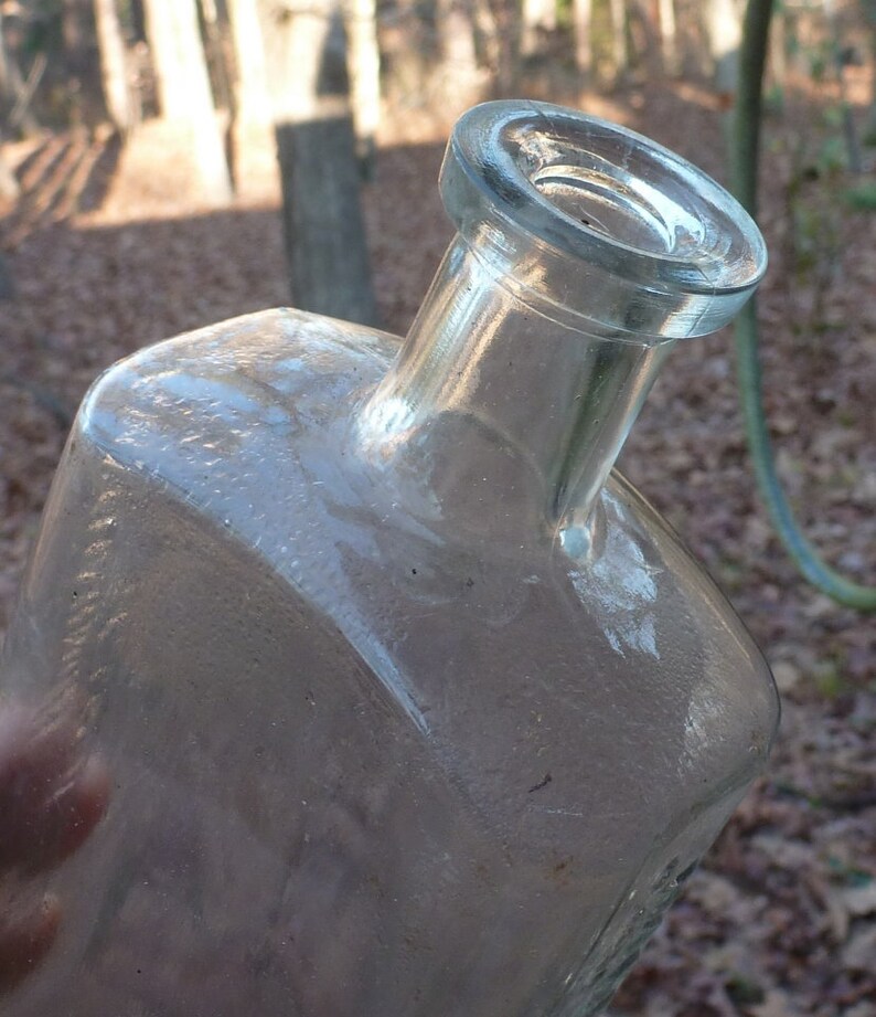 May include: A clear glass bottle with a wide mouth and a rounded, slightly flared top. The bottle has a textured surface and appears to be old.