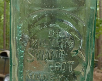 May include: A vintage, rectangular, light green glass bottle. The bottle has embossed text that reads "Dr. Kilmer's Swamp-Root Kidney, Liver & Bladder Remedy". The bottle is empty and has a rounded neck.