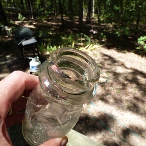 Op de afbeelding: Een heldere glazen pot met een brede, schroefdraadmond. De pot wordt in een hand gehouden, met een vage buitenachtergrond van bomen en zonlicht.