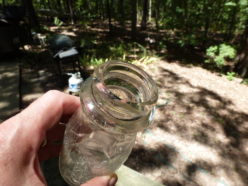 Op de afbeelding: Een heldere glazen pot met een brede, schroefdraadmond. De pot wordt in een hand gehouden, met een vage buitenachtergrond van bomen en zonlicht.