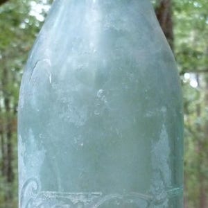 May include: An antique Coca-Cola bottle in aqua-coloured glass, featuring embossed lettering. The bottle has a traditional shape with a slender neck and a broader base. The glass has a slightly frosted look, indicating age. A collectable item.