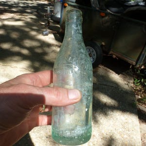 May include: A clear, vintage Coca-Cola bottle with embossed lettering. The bottle is held in a hand and appears to be made of green-tinted glass. The bottle is cylindrical with a rounded base and a narrow neck.
