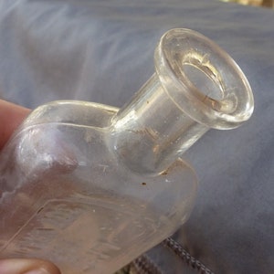 May include: A clear glass bottle with a wide mouth and a rounded body. The bottle has a textured surface and appears to be old.