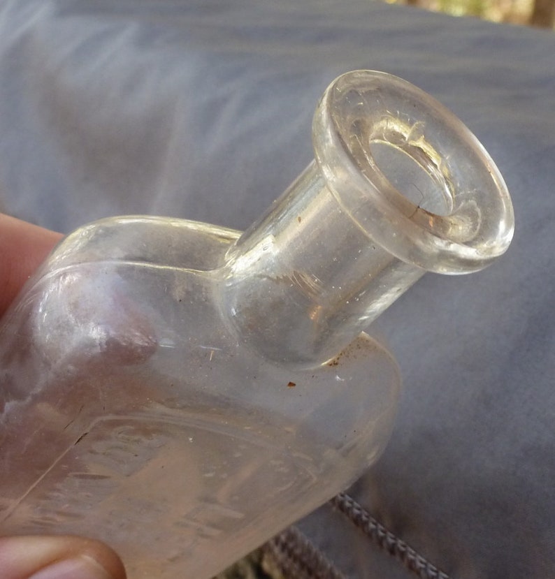 May include: A clear glass bottle with a wide mouth and a rounded body. The bottle has a textured surface and appears to be old.