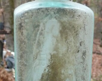 Puede incluir: Una botella de vidrio azul claro, de forma rectangular y antigua. Tiene un cuello largo y una base ligeramente redondeada. La superficie está cubierta de suciedad y mugre, lo que le da un aspecto envejecido. La botella es probablemente vintage.